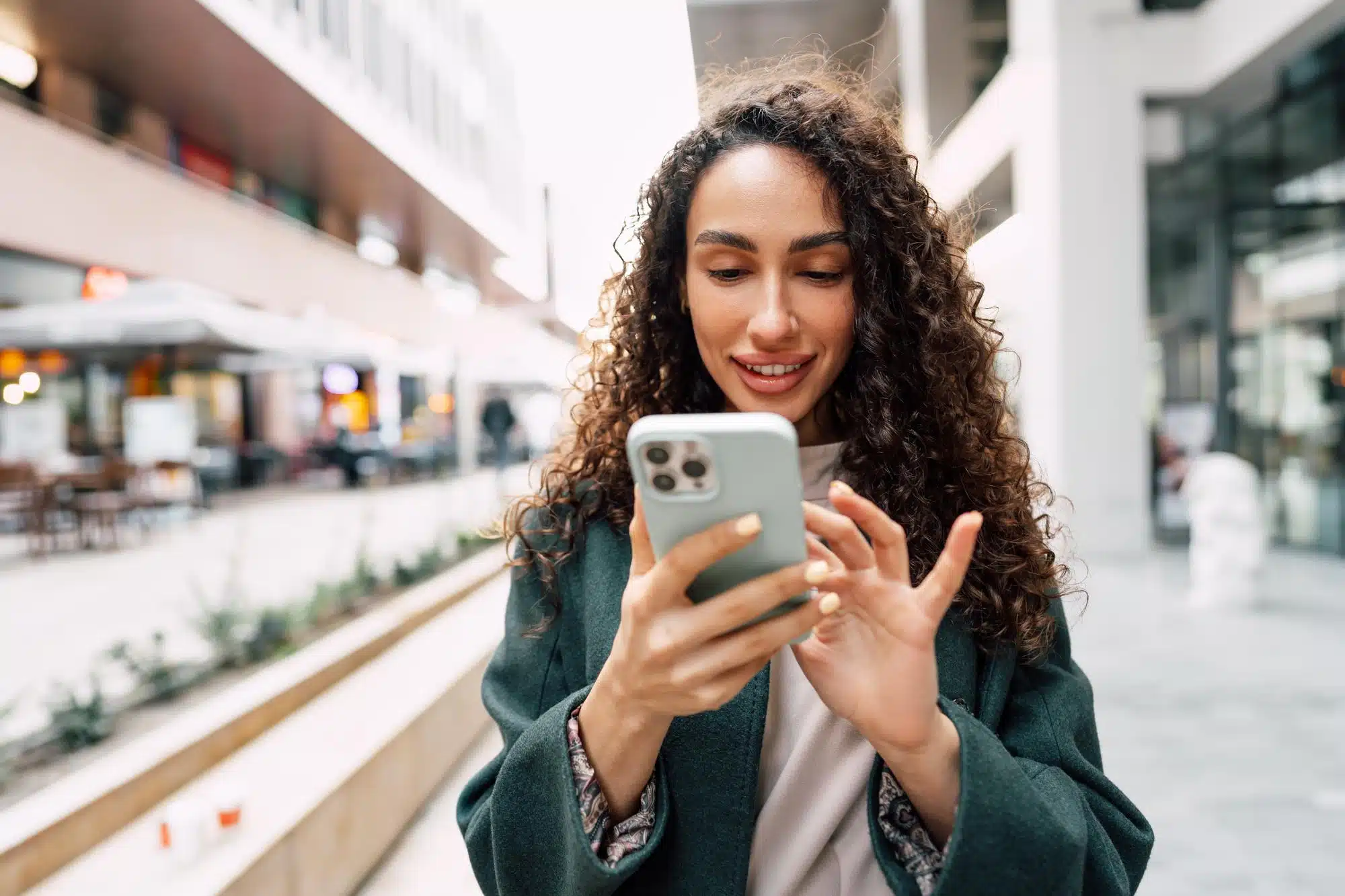 Mulher sorrindo enquanto usa iPhone Pro Max branco em rua de shopping.
